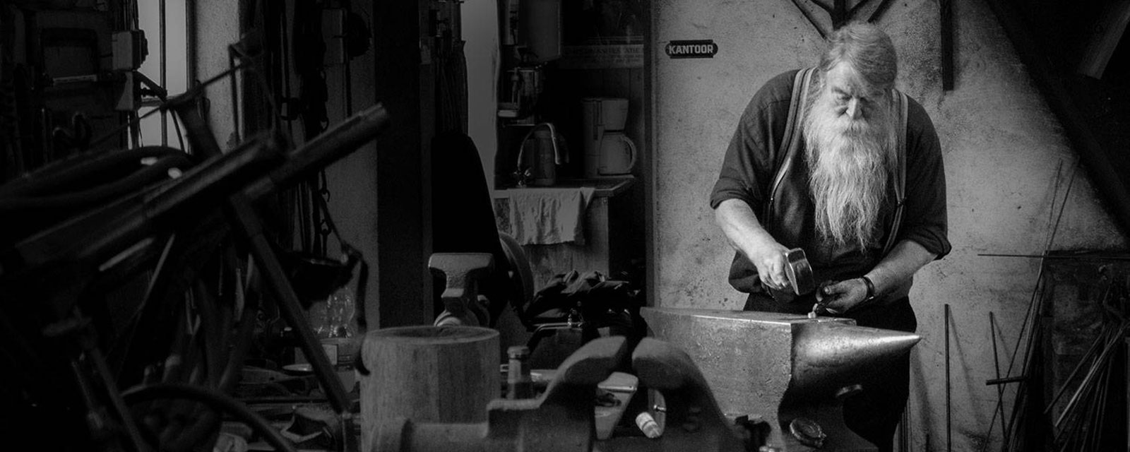 A bearded man working at an anvil in his workshop